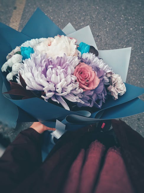 A vibrant bouquet of fresh flowers arranged with a mix of large white and pale purple chrysanthemums, soft pink roses, and smaller blue and white blossoms, all wrapped in elegant blue and white paper.