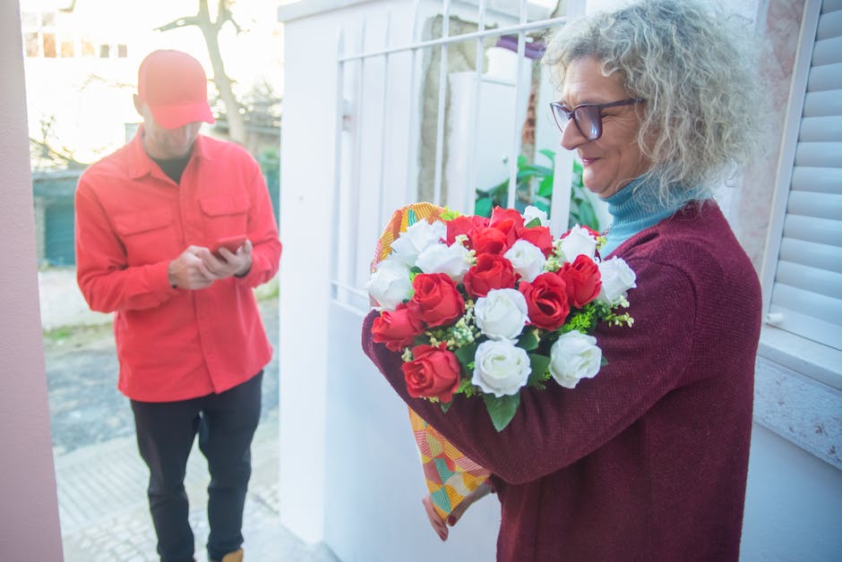 A woman with curly gray hair and glasses is holding a vibrant floral bouquet featuring red and white roses, wrapped in colorful patterned paper, during a flower delivery. She stands near a white windo