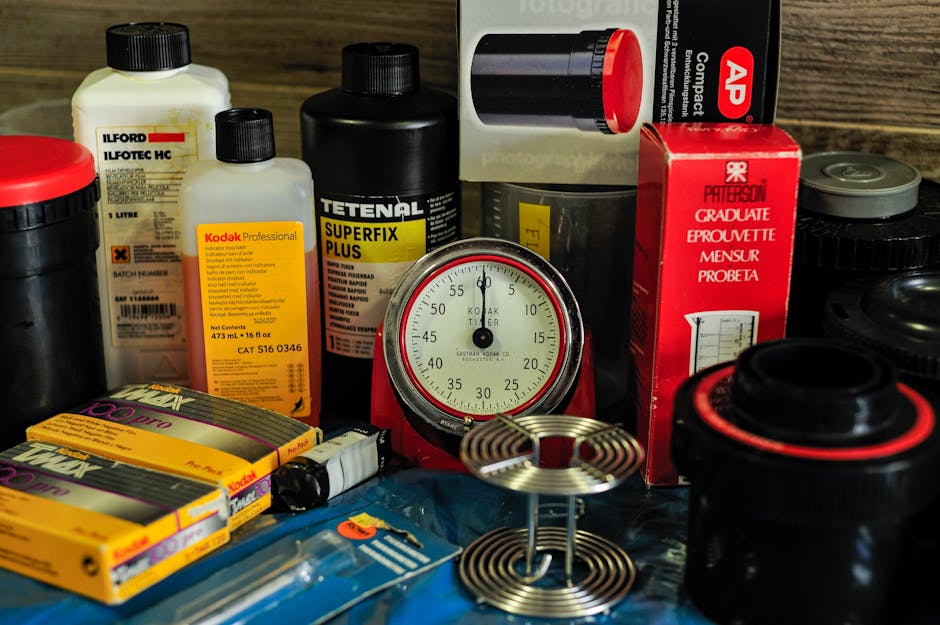 A close-up view of various photographic chemicals and equipment, including bottles of Ilford chemicals, Kodak developer, a black container, and film rolls, arranged on a wooden surface with a vintage