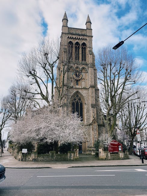 This image depicts a tall, historic stone church with Gothic architectural features, including pointed arches and a large clock face on the front facade. Surrounding the church are leafless trees tran