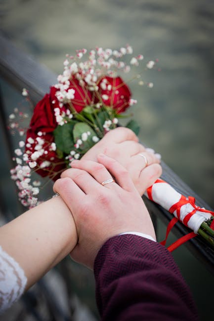 A close-up of a bride and groom's hands clasped together over a dark railing, with the bride holding a bouquet of vibrant red roses accented by delicate white baby's breath and lush green foliage. The