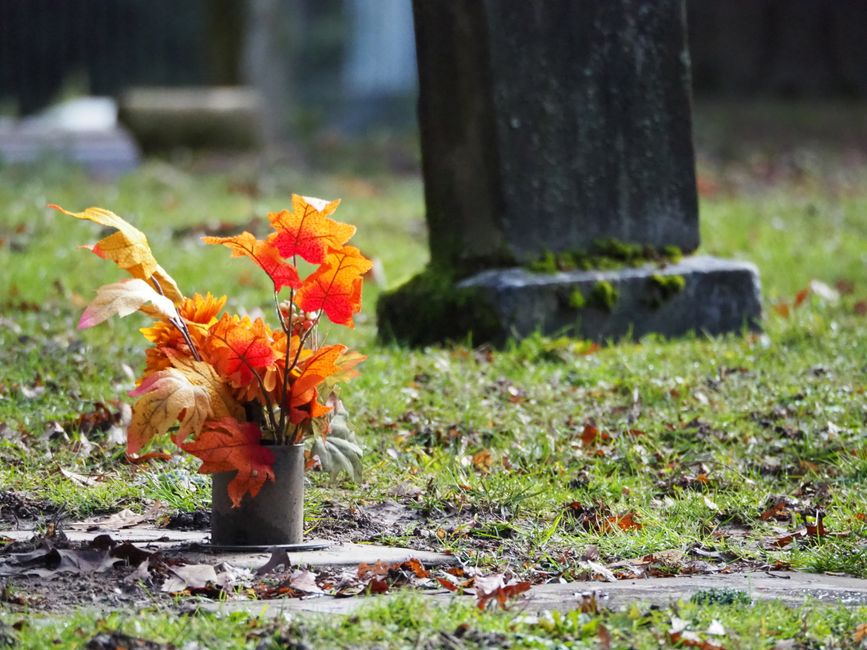 A bouquet arrangement of artificial autumn leaves in shades of orange, red, yellow, and beige, placed in a small gray cylindrical vase on a green grassy area with fallen leaves. The background feature