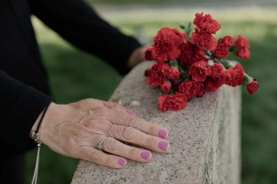 A person's left hand, adorned with a diamond ring and a black bracelet, rests gently on a concrete memorial bench. Resting atop the bench is a bouquet of vibrant red carnations, arranged simply with s