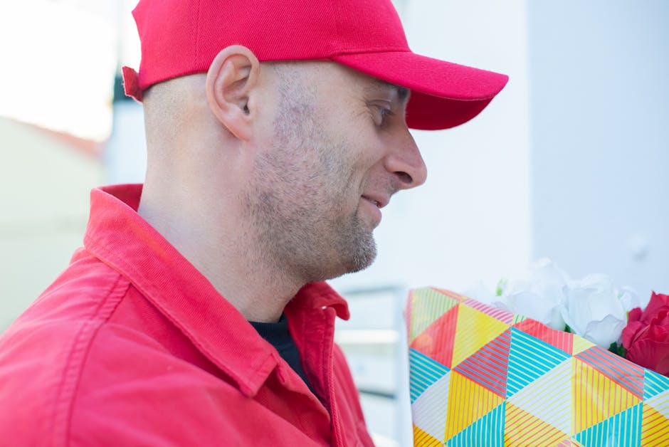 A delivery person wearing a red cap and matching red jacket is holding a vibrant floral bouquet wrapped in multicolored geometric-patterned paper. The bouquet contains an assortment of fresh flowers,