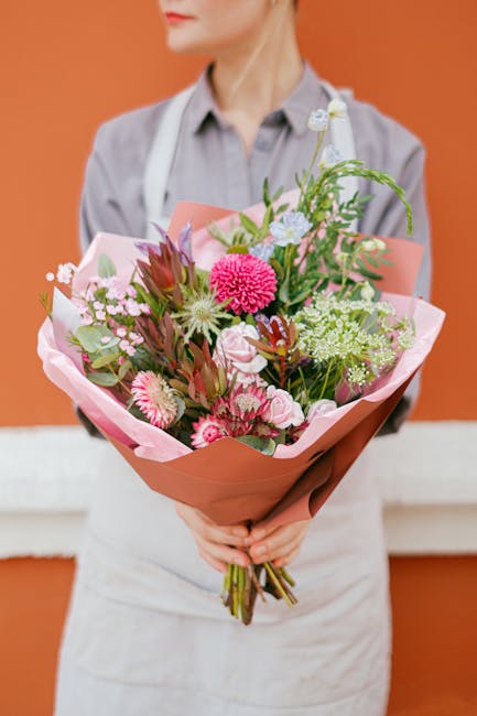 A person dressed in a light grey shirt and white apron holding a vibrant bouquet of fresh flowers, including pink and white chrysanthemums, green foliage, and purple-tipped leaves, wrapped in light pi